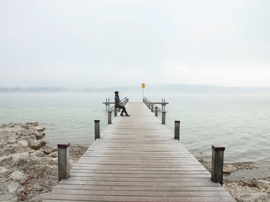Nebelverhangener Bodensee mit einem langen Steg, auf dem eine Person auf einem Pfahl sitzt.
