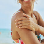 Frau cremt sich am Strand die Schulter mit Sonnencreme ein